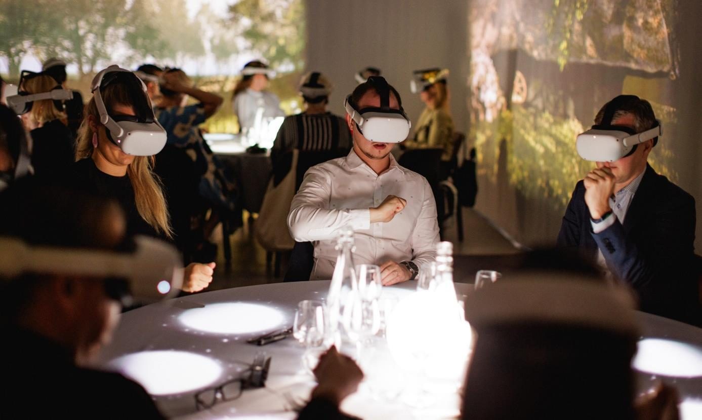 Mensen aan een ronde, verlichte dinertafel dragen witte VR-brillen, met projecties van bomen op de wand erachter.