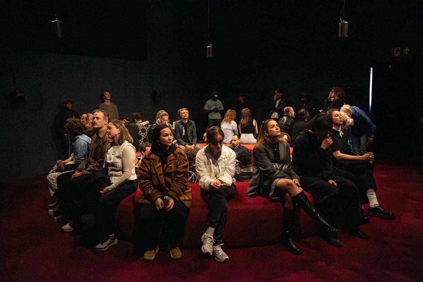 Publiek zit dicht bij elkaar op een rond rood podium in een donkere zaal met luidsprekers die aan het plafond hangen.